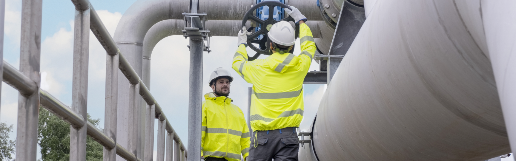 Due operatori con giacche ad alta visibilità e caschetti di protezione regolano la valvola di una tubazione in un impianto industriale di Acea Ambiente