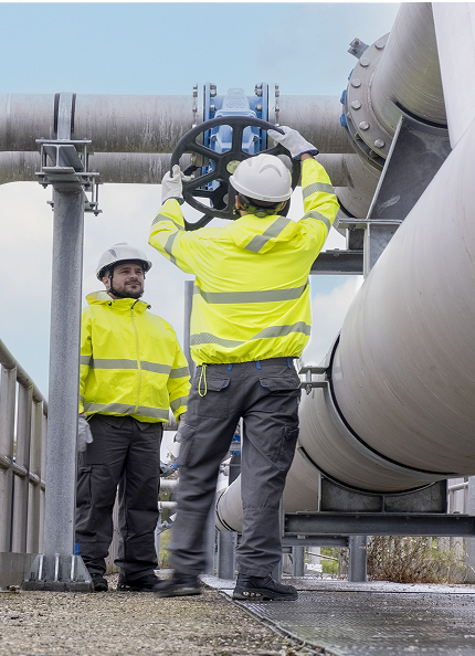 Due operatori con giacche ad alta visibilità e caschetti di protezione regolano la valvola di una tubazione in un impianto industriale di Acea Ambiente